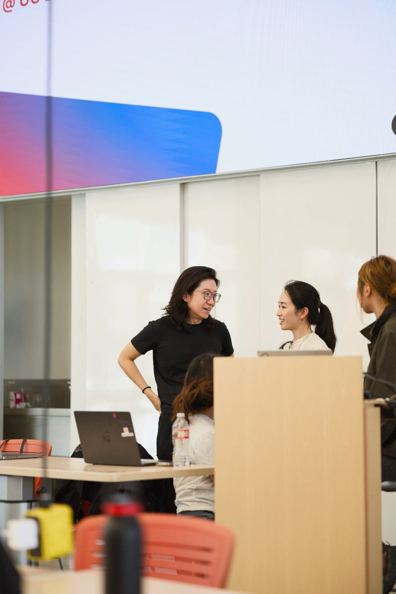 People talk at the front of the room under the big screen between demos